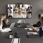 Employees speaking online to diverse freelancers on wall display and office workers at meeting table