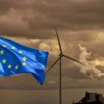 Waving flag of Europe in front of a polluting factory chimney with smoke and wind turbine
